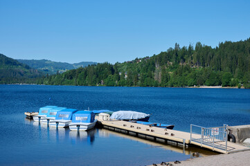 Fototapeta premium Titisee with blue water and boats on the jetty, blue sky, mountains and green trees