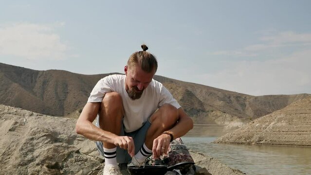 A handsome young male hiker in a hike prepares food on the seashore using a frying pan and a portable burner with interchangeable gas cylinders. Frying sausages on a trip.