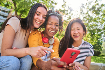 Three happy women using cell phone outdoors. Group of smiling female friends viewing social...