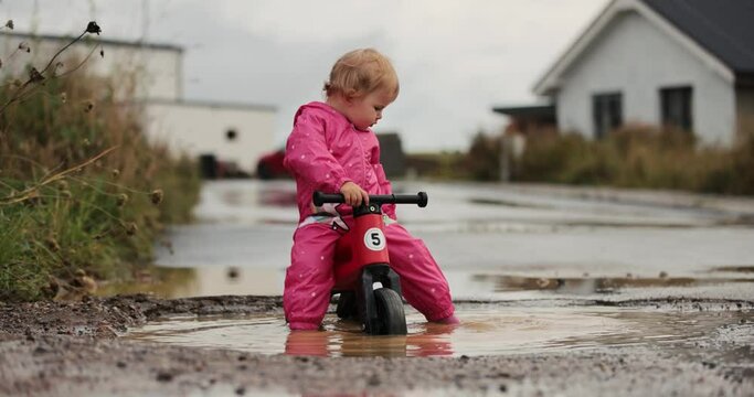 Small oneyear old baby girl  and mother playing in mud puddles after heavy rain. Jumping, throwing rocks and examine new . Wearing pink purple raincoat and Wellingtons. Riding a red bike, dogs around