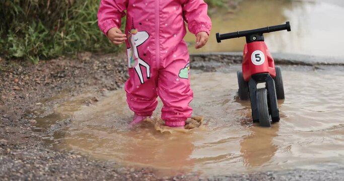 Small oneyear old baby girl  and mother playing in mud puddles after heavy rain. Jumping, throwing rocks and examine new . Wearing pink purple raincoat and Wellingtons. Riding a red bike, dogs around