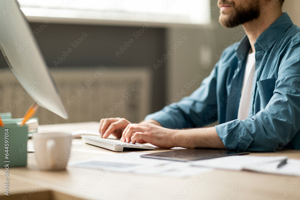 Poster bearded young man sitting at desk and typing on computer keyboard
