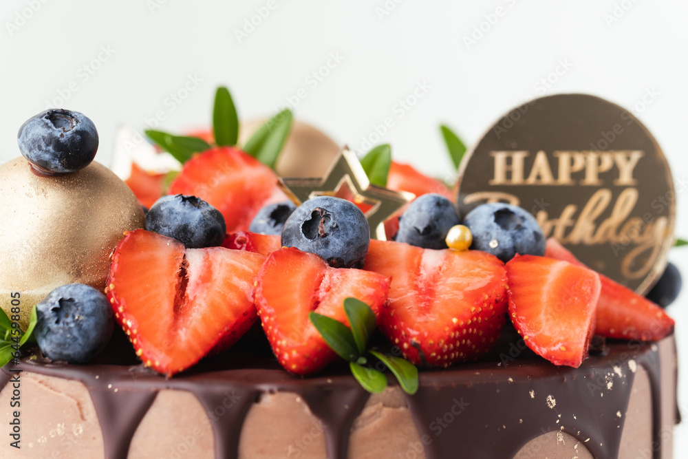 Wall mural Birthday cake with milk chocolate frosting decorated with melted dark chocolate drips and fresh summer berries: strawberries and blueberries on top. White background