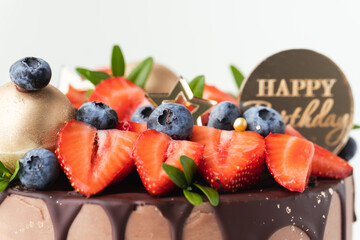 Birthday cake with milk chocolate frosting decorated with melted dark chocolate drips and fresh summer berries: strawberries and blueberries on top. White background