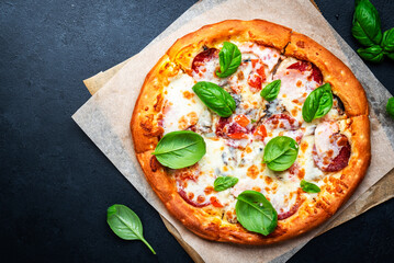 Homemade pizza with italian salami sausage, mozzarella cheese, tomato sauce and green basil on black kitchen table background, top view