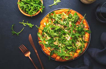 Hot pizza with mozzarella cheese, ham, pesto and fresh arugula, black table background, top view