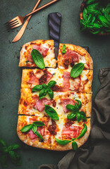 Hot Roman pizza with ham, pancetta, mozzarella cheese, sun dried tomatoes, sauce and green basil, rustic green table background, top view - obrazy, fototapety, plakaty