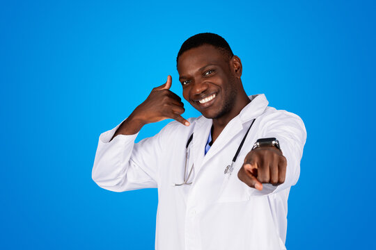 Cheerful young african american man doctor in white coat pointing finger at camera and sign call me with hand