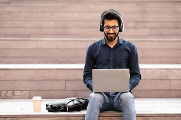 Concentrated indian guy freelancer using laptop, headphones outdoor