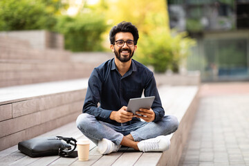 Indian guy sitting outdoor with digital tablet, having coffee break