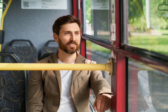 Happy brunet man sitting near window in city bus, looking thoughtfully away while riding. Portrait of bearded male passenger looking out window, while commuting to work in morning. Transport concept.