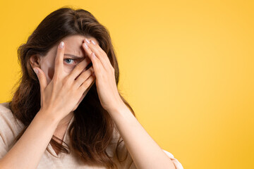 Scared young caucasian lady covers her face with hands, afraid, isolated on yellow studio background