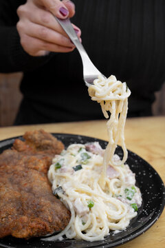 Eating at the restaurant. Closeup view of a woman's hand holding a fork, eating milanesa, breaded steak, and spaghetti with a bacon and leek creamy sauce.