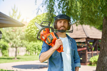 Attractive male worker in safety shield posing, while carrying rechargeable chain saw on shoulder at sunny day. Portrait of strong bearded handyman with modern electric saw. Concept of craftsmanship. 