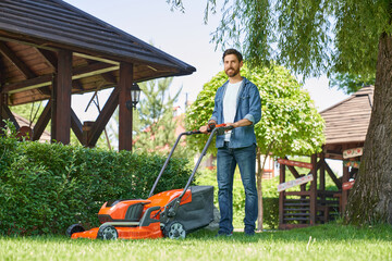 Smiling male landscaper in denim shirt trimming overgrown lawn with lawn mover at summer day. Low...