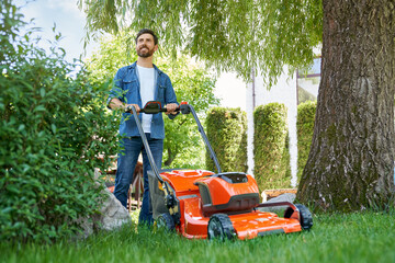 Handsome bearded man in blue shirt trimming lawn with electric mower on backyard. Low angle view of...