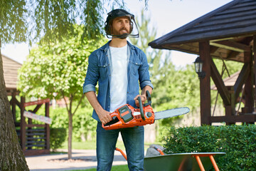 Male gardener in protective shield standing with cordless chain saw, working in summer garden. Front view of worker with beard holding modern electric saw, while looking ahead. Concept of gardening. 