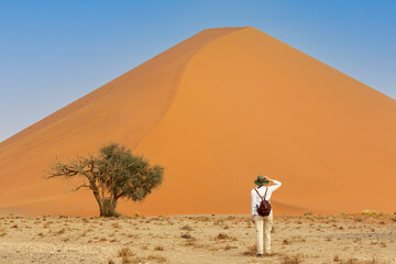 Young caucasic traveler standing  near orange sand dune with her hand on the hat. Travel desert...