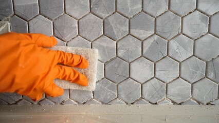 A person using a sponge to clean tiles. The final stage of laying mosaic tiles. Close-up: a gloved...