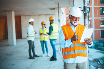 Mature man working on digital tablet at construction site. Mid adult manual worker with blue hardhat inspecting construction site while checking list on laptop.