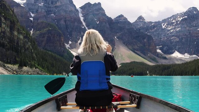 Canoeing on the bright blue glacial water of Moraine Lake with tall jagged mountains and dense pine forests, Banff National Park, Alberta, Canada. Traveller paddling a canoe.