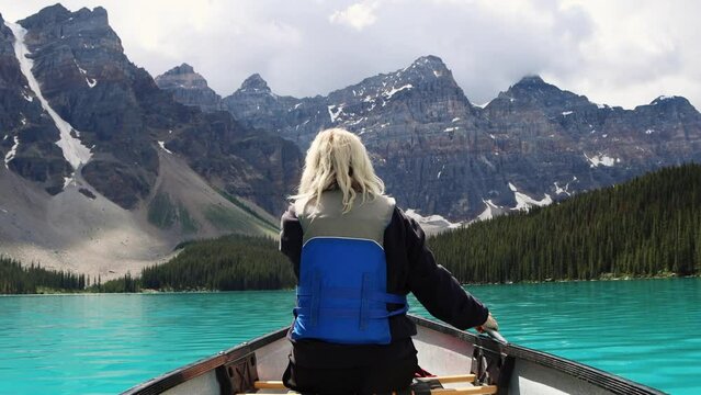 Canoeing on the bright blue glacial water of Moraine Lake with tall jagged mountains and dense pine forests, Banff National Park, Alberta, Canada. Traveller paddling a canoe.