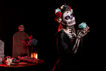 Spooky santa muerte model posing with roses over black background, looking like goddess of death. Beautiful woman with skull make up and body art, festival holiday celebration in studio.