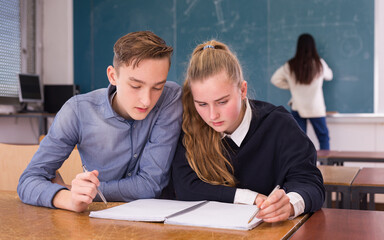 Intelligent teenager helping girl classmate prepare for exam, explaining study material in college classroom
