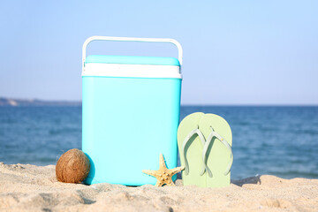 Modern beach cooler, flip-flops and ripe coconut on sand