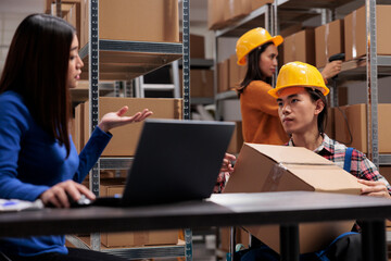 Asian warehouse employee holding cardboard box and making inventory with coworker. Delivery manager checking order pick ticket on laptop while sitting at desk in industrial storehouse