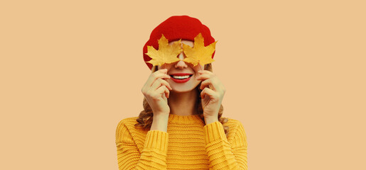 Autumn color style outfit, portrait of beautiful smiling young woman covering her eyes with yellow maple leaves wearing red french beret hat, knitted sweater on brown studio background