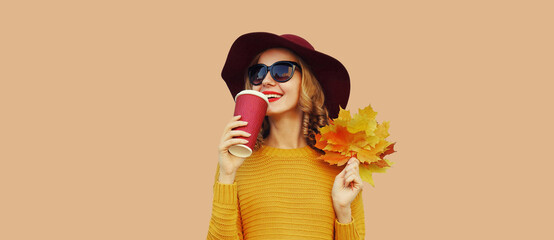 Autumn color style outfit, portrait of beautiful smiling young woman with yellow maple leaves drinks coffee wearing hat, knitted sweater on brown studio background