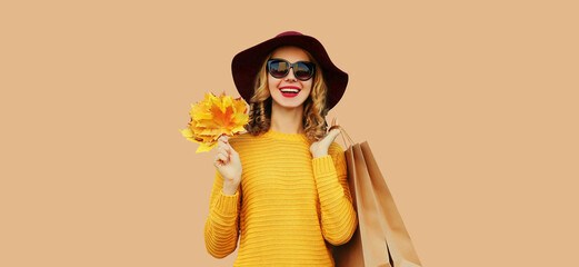 Autumn color style outfit, portrait of beautiful young woman with shopping bag holds yellow maple leaves wearing hat, knitted sweater on beige studio background