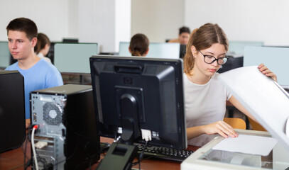 Teenage girl sitting at table and using scanner in computer class in school.