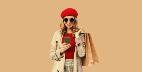 Autumn style outfit, portrait of stylish smiling young woman model with mobile phone holds shopping bags wearing red french beret hat, coat jacket, round sunglasses on beige studio background
