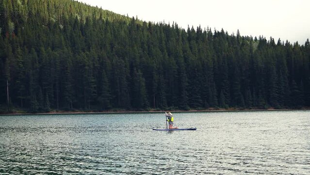 Girl paddle boarding across the emerald green water of Two Jack Lake, Banff national park, Alberta, Canada. Surrounded by tall mountains and dense green forests.