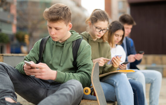 Portrait of boy teenager addicted in phone sitting with friends on bench outdoors
