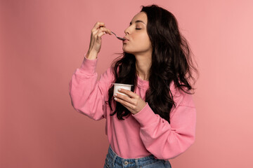 Waist-up portrait of a young woman snacking on the yogurt with a spoon from the plastic cup during...