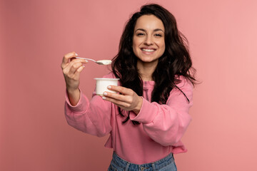 Waist-up portrait of a joyous brunette snacking on her favorite yogurt with the teaspoon from the plastic cup during the studio photo shoot
