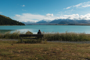 A man sitting beside a blue lake with snow capped mountains in the background.