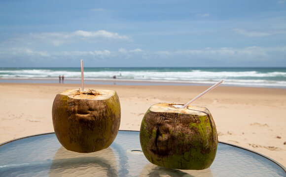 Cocos com canudinho e cadeira de praia vazia em praia ensolarada do nordeste brasileiro, com pessoas distantes em trajes de banho nas ondas do mar