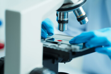 Close up dedicated hematology lab technician scrutinizing blood sample analysis under microscope, scientific laboratory research and drug testing concept, red color liquid