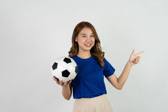 Happy Asian female soccer fan sending support to favorite team with soccer ball, smiling woman in blue t-shirt holding soccer ball to cheer at soccer game, isolated on white background.