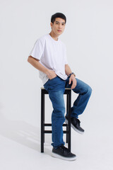 Fototapeta na wymiar Portrait isolated cutout studio shot Asian handsome cool male fashion model in casual fashionable t shirt and blue jeans sitting on chair holding hand in pocket look at camera on white background