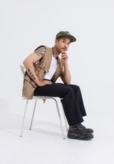 Fototapeta premium Portrait isolated cutout studio full body shot Asian vintage classy mustache neck arms tattoos male fashion model in casual fashionable safari vest and cap sitting on white chair on white background.
