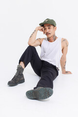 Isolated cutout studio shot Asian vintage classy mustache with neck arms hands tattoos male fashion model in casual fashionable sleeveless shirt cap boots sitting on floor on white background.