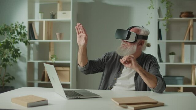 A Modern Old Man in VR Glasses Examines and Touches Everything Around. An Elderly Man in VR Glasses Sitting at a Table with a Laptop.Virtual Reality Games for the Elderly.
