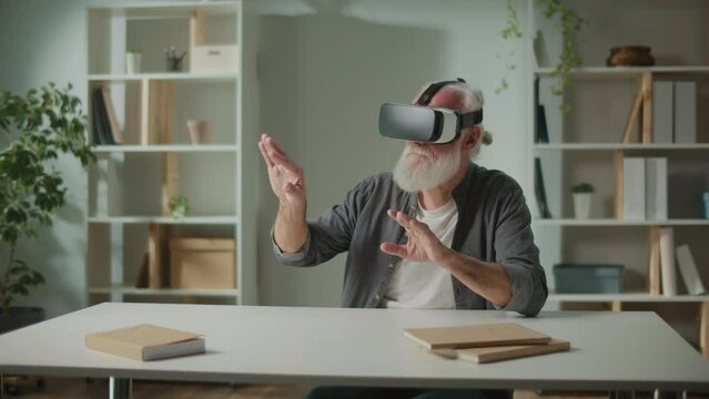 A Modern Old Man in VR Glasses Examines and Touches Everything Around. An Elderly Man in VR Glasses Sitting at a Table at Home.Virtual Reality Games for the Elderly.