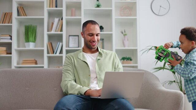 Happy multiethnic man in casual clothes working on laptop while two boys running around sofa with toy guns. Calm stay-at-home parent performing task for employer while keeping kids entertained.