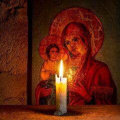 A lit candle against the background of an ancient religious icon. A religious icon and a candle on a rough wooden shelf.
